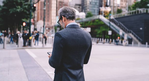Back view of stylish gray haired mature businessman in black costume and glasses looking at mobile on blurred urban background in New Yor - Foto, Imagem