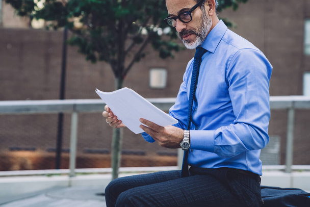 Side view of middle aged man in business suit and glasses sitting on bench and analyzing papers on New York street - Foto, afbeelding
