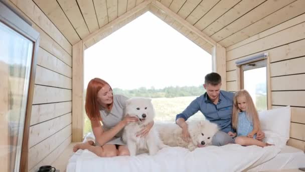 Young Caucasian family couple with baby daughter in a small modern rustic house with a large window. Lying on the bed, hugging, playing and looking out the window. Two Samoyeds. Weekend vacation - Footage, Video