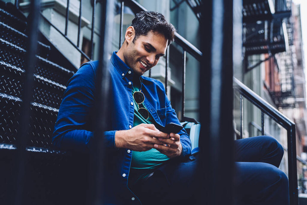 Young happy trendy man in blue shirt chilling on metal porch on street and browsing smartphone smilingly, New York city - Фото, изображение