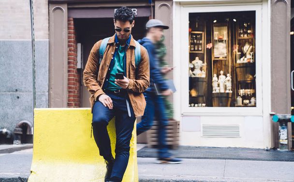 Serious young stylish male in sunglasses and casual clothes surfing internet and texting in modern digital smartphone while sitting and looking down at street in city - 写真・画像
