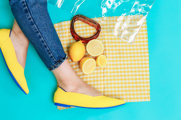 beautiful female legs are dressed in stylish yellow shoes without a heel. Light yellow summer sandals on a blue background against the background of a handbag and bright lemons. - Foto, afbeelding