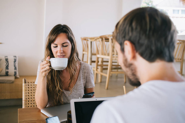 Young woman with brown hair and smartphone drinking coffee from white cup and sitting at wooden desk near bearded man working on laptop in cafe - Foto, Imagem