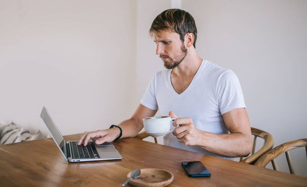 Focused casual man in white t-shirt having coffee while sitting at table with smartphone and surfing laptop working remotely in cafe - Foto, afbeelding