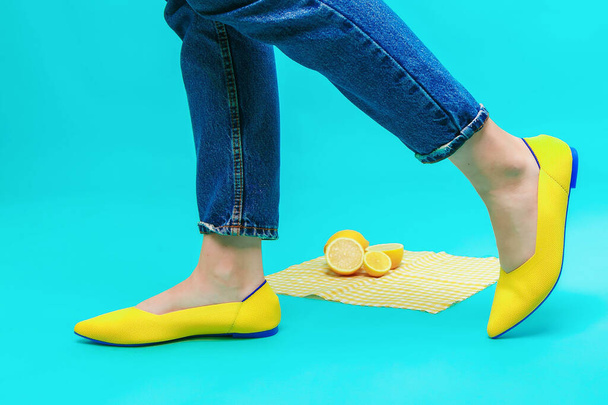 美しい女性の足はヒールのないスタイリッシュな黄色の靴を履いています。明るいレモンの背景に青の背景に薄い黄色の夏のサンダル. - 写真・画像
