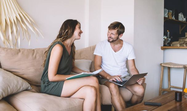 Hombre guapo alegre navegando portátil y mujer feliz con cuaderno en las manos sentados en el sofá en casa trabajando mirándose el uno al otro
 - Foto, imagen