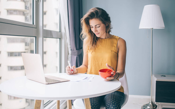 Giovane donna attraente concentrata in camicetta gialla senza maniche seduta al tavolo rotondo bianco con laptop e tazza rossa che scrive su carta a casa
 - Foto, immagini