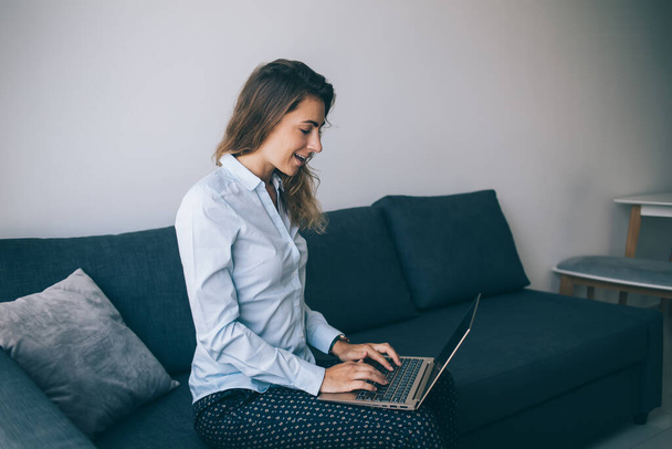 Modern adult smiling woman with wavy hair working on laptop while sitting on sofa in living room of modern apartment  - Φωτογραφία, εικόνα
