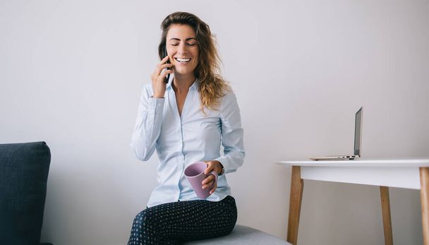 Young female employee sitting with eyes closed holding coffee cup and smiling while talking on phone in office against white background - Photo, Image