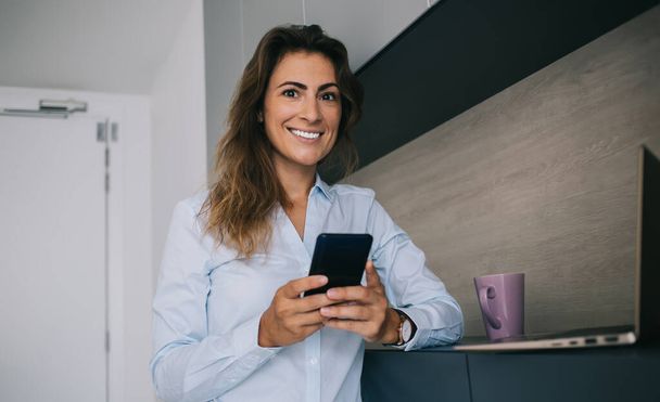 Pretty optimistic woman in blue shirt standing with smartphone at home and looking at camera while smiling after reading good message - Foto, Imagem