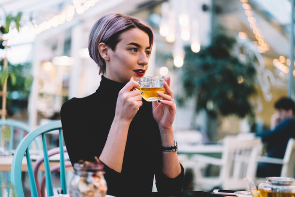 Stylish positive woman with short pink hair in casual clothes drinking cup of hot tea while sitting at table at hipster cafeteria - Foto, afbeelding