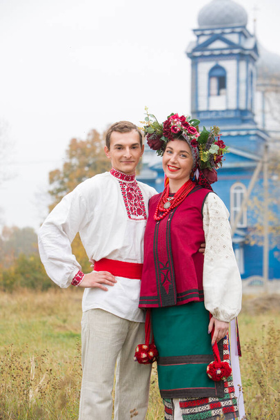 Girl and guy in retro costumes on the street in the old village. Retro staging of an ancient rite. Beautiful wreath on a girl. The guy hugs the girl, both are smiling. Antique clothing of the late - Photo, Image