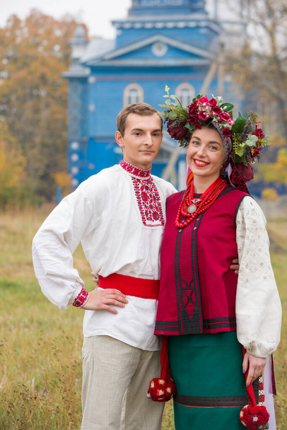 Girl and guy in retro costumes on the street in the old village. Retro staging of an ancient rite. Beautiful wreath on a girl. The guy hugs the girl, both are smiling. Antique clothing of the late - Photo, Image