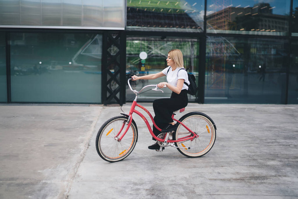 Side view of attractive young blonde woman in casual outfit with small black backpack riding pink bicycle against glass building looking away - Fotoğraf, Görsel