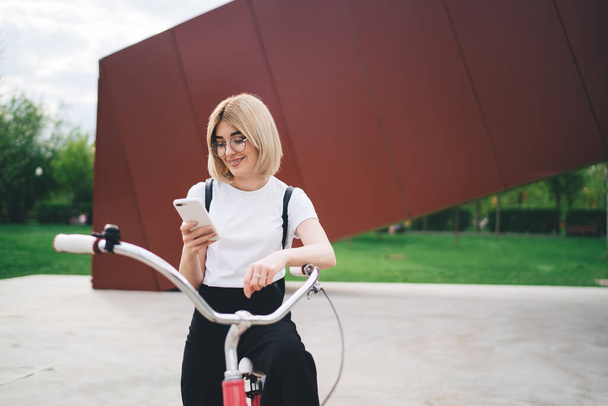 Happy female in glasses wearing casual white shirt attentively browsing smartphone with joy while sitting on bicycle on city street - Fotografie, Obrázek