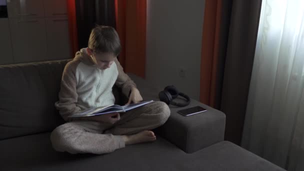 Teen boy reads a book sitting on the couch. Coronavirus epidemic 2020. - Materiał filmowy, wideo
