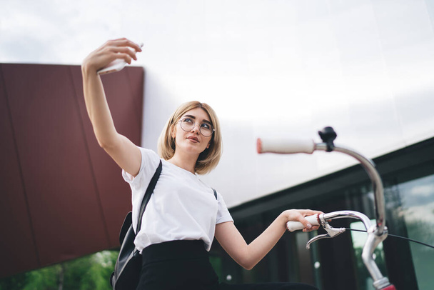 Charming young female in glasses and casual clothes taking selfie on smartphone and smiling while sitting on bike outdoors near mall - Фото, изображение