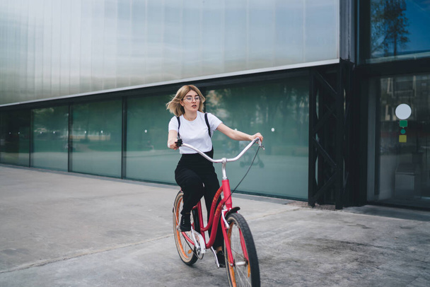 Young attractive female student in casual clothes sitting on bicycle and looking attentively frontwards while spinning pedals and cycling on street - Foto, immagini
