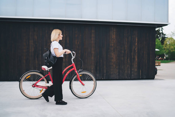 Side view of young modern blond woman in casual outfit and with black backpack walking with bicycle against street building looking away - Фото, зображення