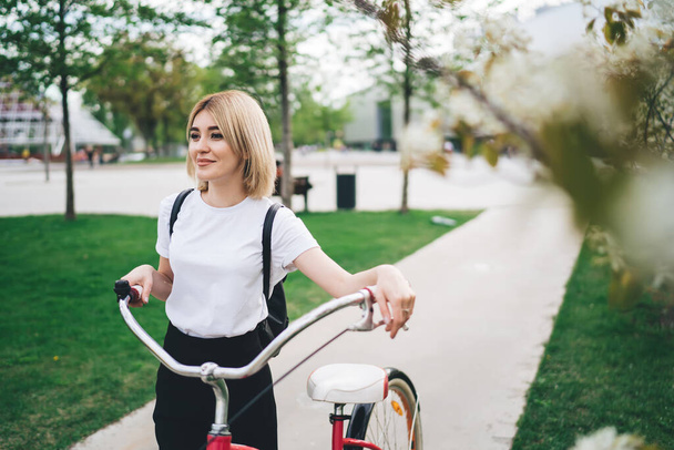 From above cheerful young blond woman in casual outfit and with black backpack walking with bicycle against blurred spring trees in sidewalk in street looking away  - Fotó, kép