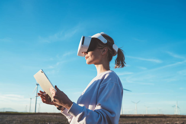 Woman in VR goggles standing turned to sunshine in wind power plant holding tablet and stylus and having virtual reality experience - Zdjęcie, obraz