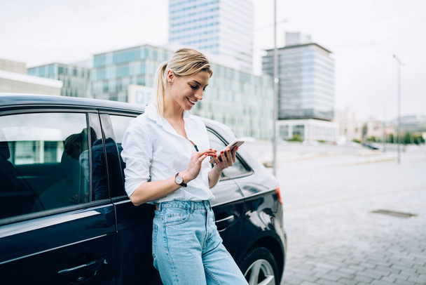 Cheerful blond female in white blouse and jeans browsing mobile phone and leaning against black car on blurred urban background - Φωτογραφία, εικόνα