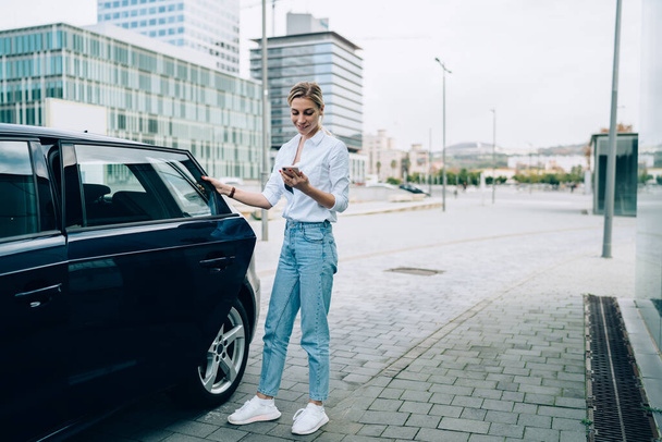 Blond slim beautiful female in white blouse and jeans browsing smartphone holding car door while standing on city street near road - Zdjęcie, obraz
