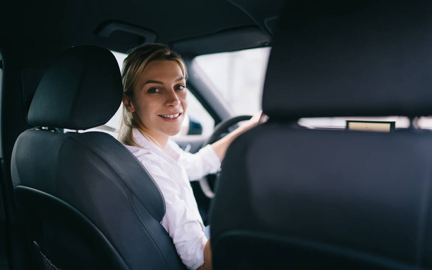 Back view of smiling blond woman in white shirt sitting at steering wheel in car and looking at camera over shoulder - Foto, immagini
