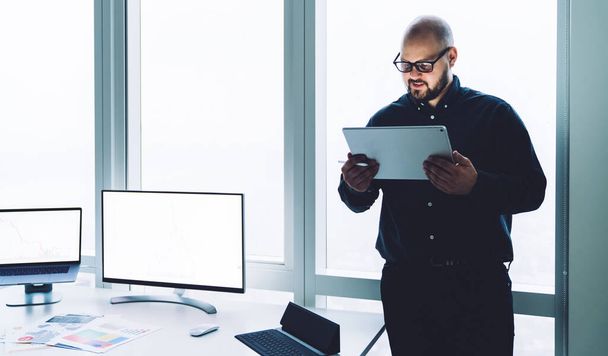 Modern bald man in glasses watching tablet while working in office standing near desk with computer and papers in modern office - Valokuva, kuva