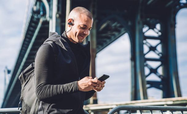 Young cheerful ethnic male in black sports clothing with backpack using phone and listening to music in city under bridge - Photo, Image