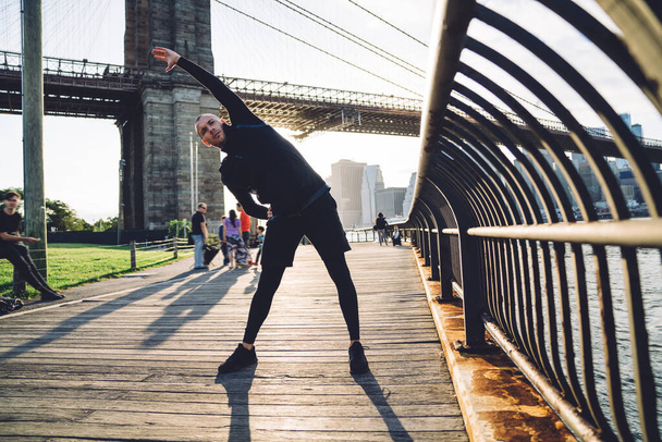 Focused sportive guy in black sportswear stretching and bending on side while standing on embankment with cityscape in backlit on background  - Foto, afbeelding