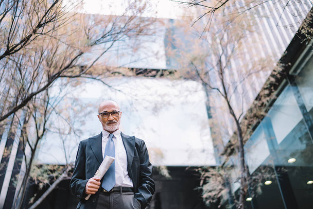 From below senior gray haired male in elegant clothes and glasses carrying newspaper in hand and looking at camera near massive building - 写真・画像