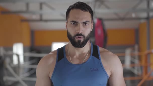 Close-up portrait of serious confident Middle Eastern boxer posing at the background of blurred boxing ring in gym. Handsome tired man looking at camera and sighing. Sport, lifestyle, endurance. - Video, Çekim