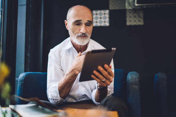 Stressed old bald man in formal clothes using modern tablet with sad expression sitting in armchair with hand on chest at table at cafe - 写真・画像
