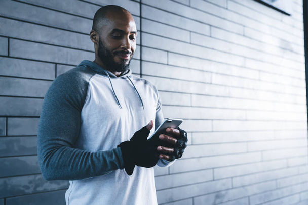 Smiling bearded adult ethnic bodybuilder in gray sweater and gloves using smartphone while working out in fitness club in front of gray brick wall  - Fotografie, Obrázek