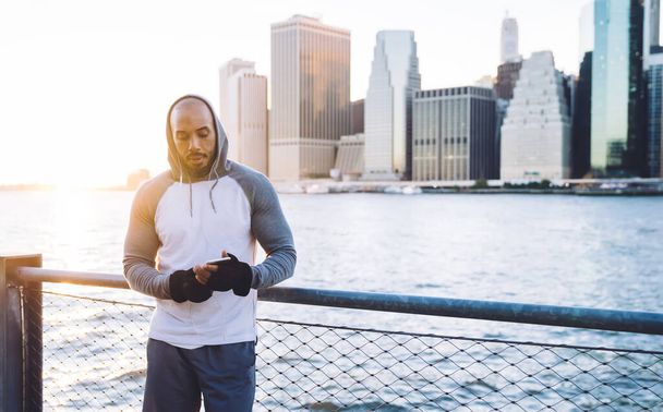 Sporty adult African American male in tracksuit using earphones and surfing smartphone while standing near fence against city skyscrapers in back lit - Photo, image