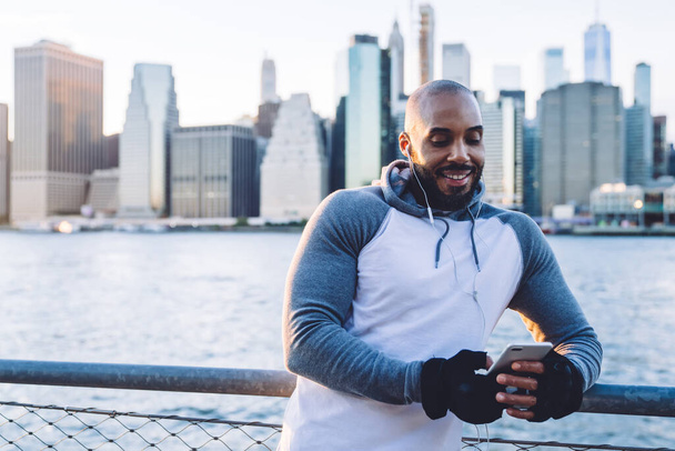 Cheerful black ethnic man in sportswear and earphones browsing smartphone while standing at fence in morning sunlight with city background  - Foto, Imagem
