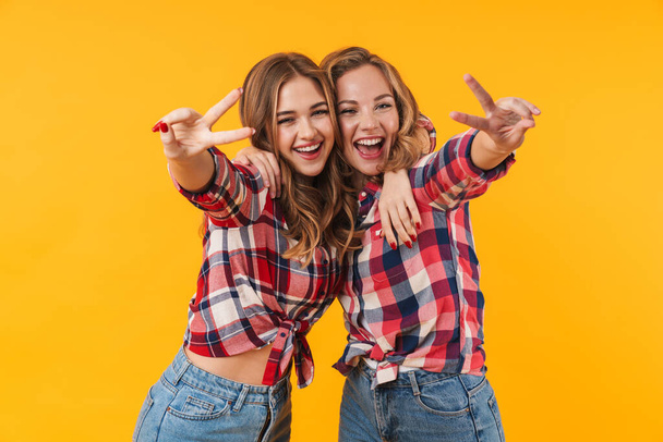 Image of two young beautiful girls wearing plaid shirt smiling and gesturing peace sign isolated over yellow background - Valokuva, kuva