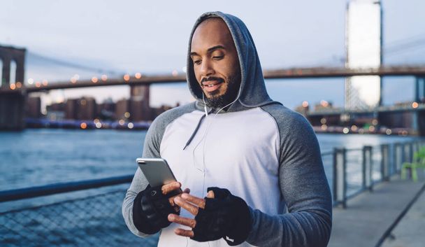 Young African American man standing and listening to music in earphones while texting on smartphone and slightly smiling against background of river and bridge - Foto, Imagem