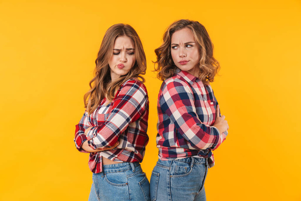 Image of two young beautiful girls wearing plaid shirt frowning and standing back to back isolated over yellow background - Zdjęcie, obraz