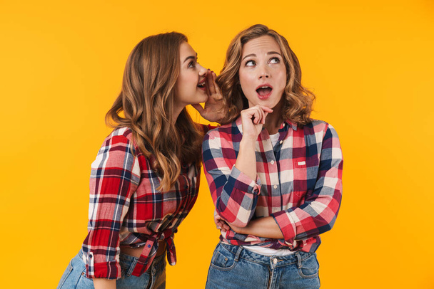 Image of two young beautiful girls wearing plaid shirt talking gossips or secrets to each other isolated over yellow background - Fotografie, Obrázek