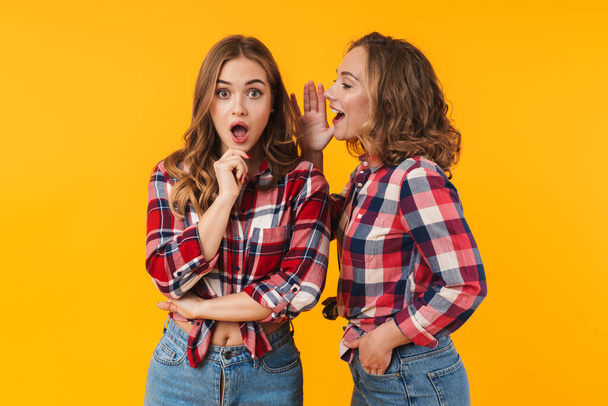 Image of two young beautiful girls wearing plaid shirt talking gossips or secrets to each other isolated over yellow background - Foto, immagini