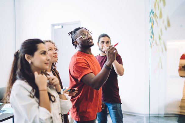 Side view of young multiracial men and women gathering in office with bright sticky notes on glass wall while planning project  - Fotografie, Obrázek