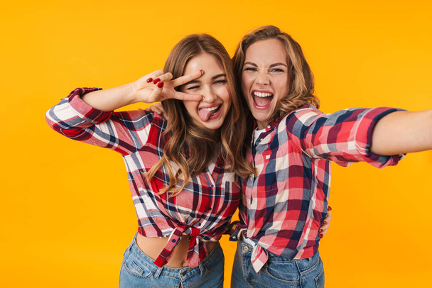 Image of two young beautiful girls wearing plaid shirt smiling and hugging while taking selfie photo isolated over yellow background - Fotografie, Obrázek