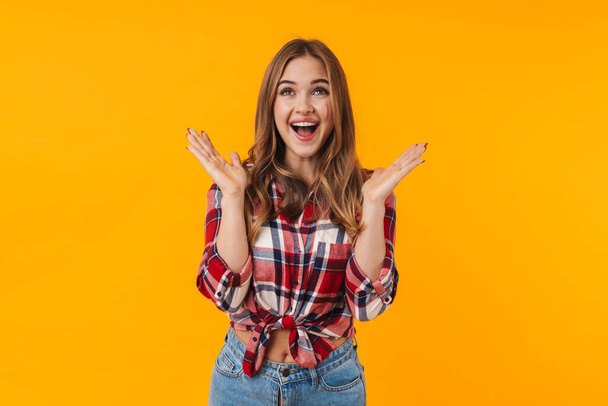Image of happy young attractive woman wearing plaid shirt screamng and raising hands isolated over yellow background - Zdjęcie, obraz