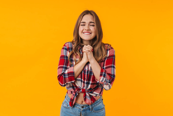 Image of happy young attractive woman wearing plaid shirt praying or making wish isolated over yellow background - Fotoğraf, Görsel