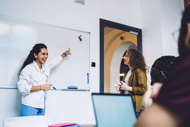 Casual young cheerful woman pointing on blank whiteboard while talking to university friends coworking on modern project in study room  - Fotografie, Obrázek