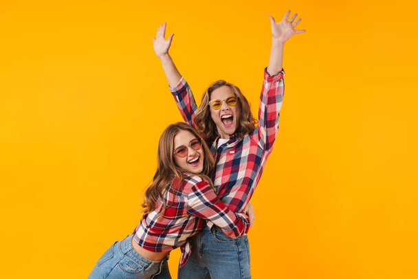 Image of two young beautiful girls wearing plaid shirts smiling and having fun isolated over yellow background - Zdjęcie, obraz
