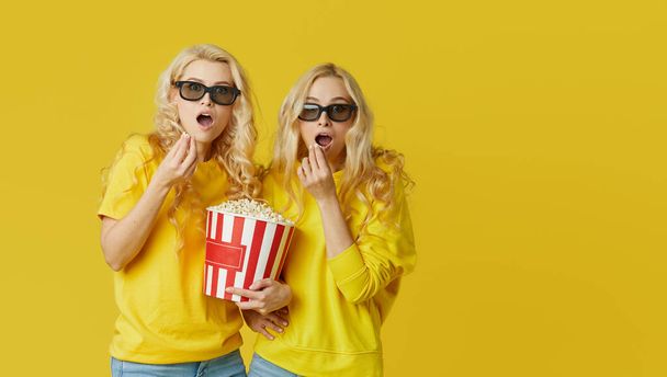 Shocked young models women in 3d glasses eating popcorn, looks scary Movie at the cinema. Isolated on yellow background - Fotografie, Obrázek