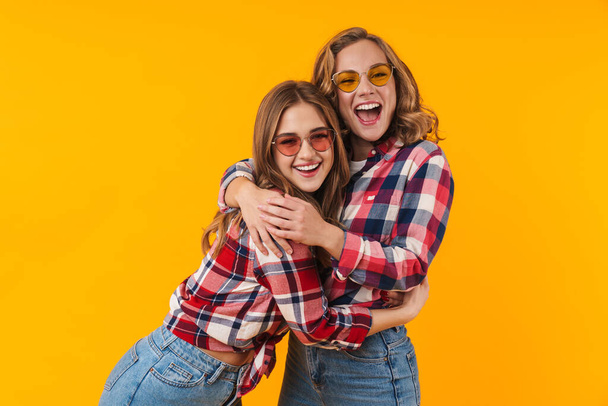 Image of two young beautiful girls wearing plaid shirts smiling and having fun isolated over yellow background - Valokuva, kuva